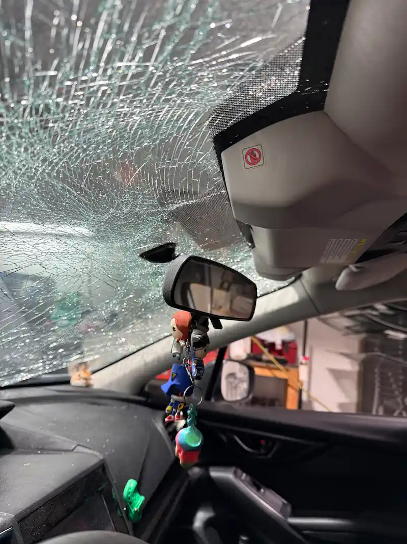 Interior view of a Subaru Impreza with the windshield shattered by a hood strike at highway speed — glass still intact in the frame but completely crazed.
